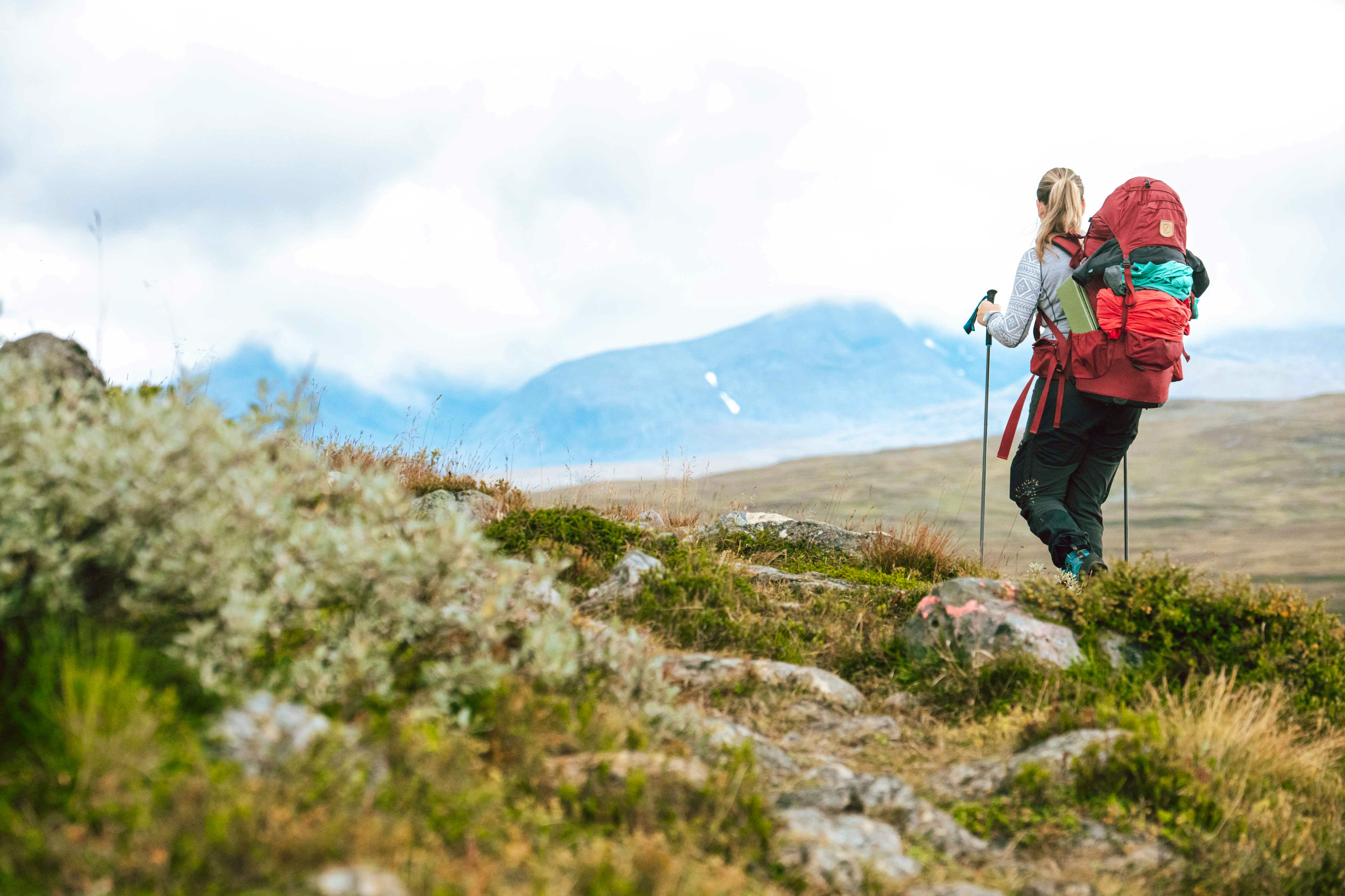 A woman with a large backpack and trekking poles hikes uphill in the mountains, with a wide view of the surrounding landscape.