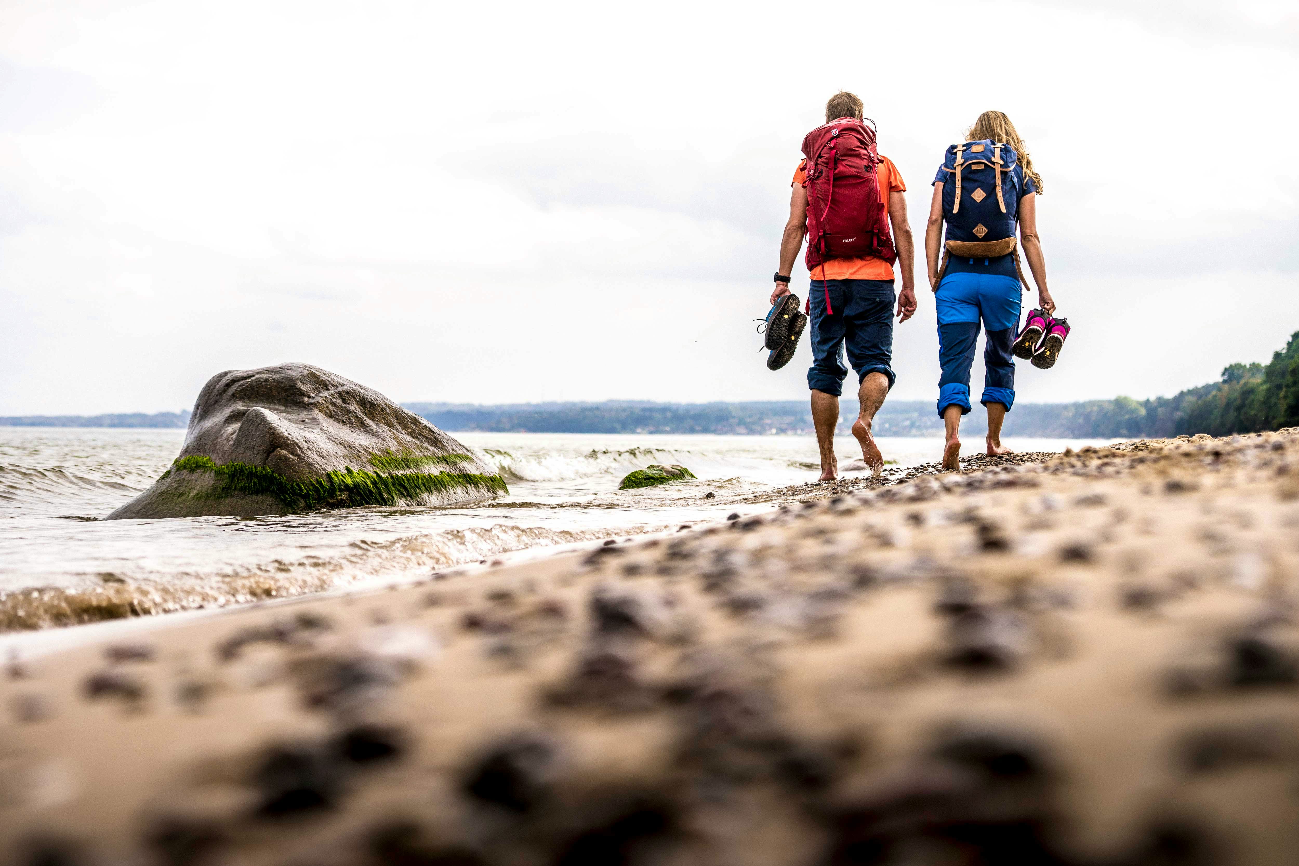 Two hikers walk barefoot along a sandy beach, carrying their shoes and backpacks, with the sea and rocks in the background.