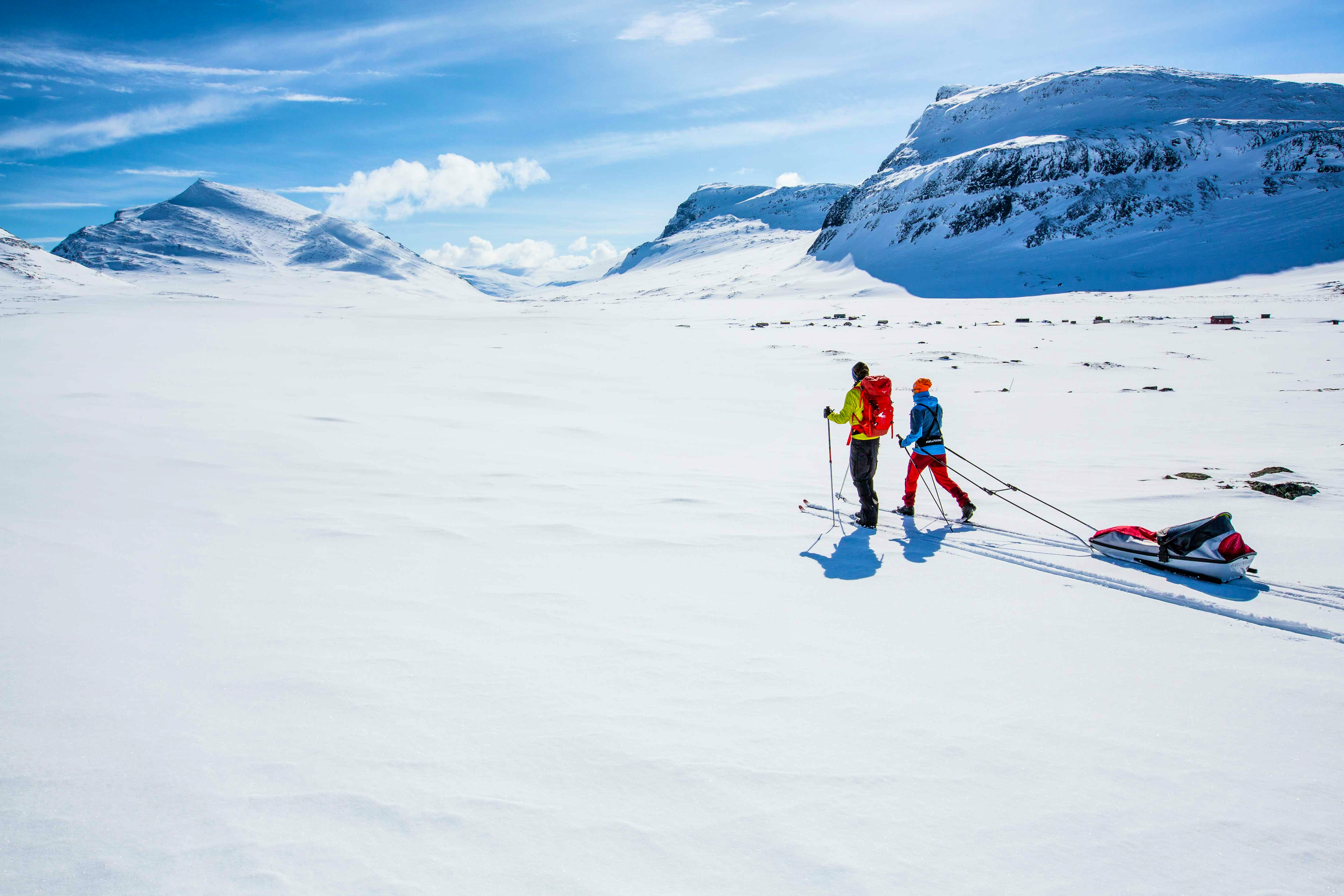 Two people on backcountry skis pull sleds across a snow-covered mountain landscape under a clear blue sky.