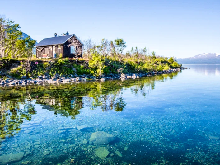 Sauna by the shore of Lake Torneträsk with clear water and mountains in the background.
