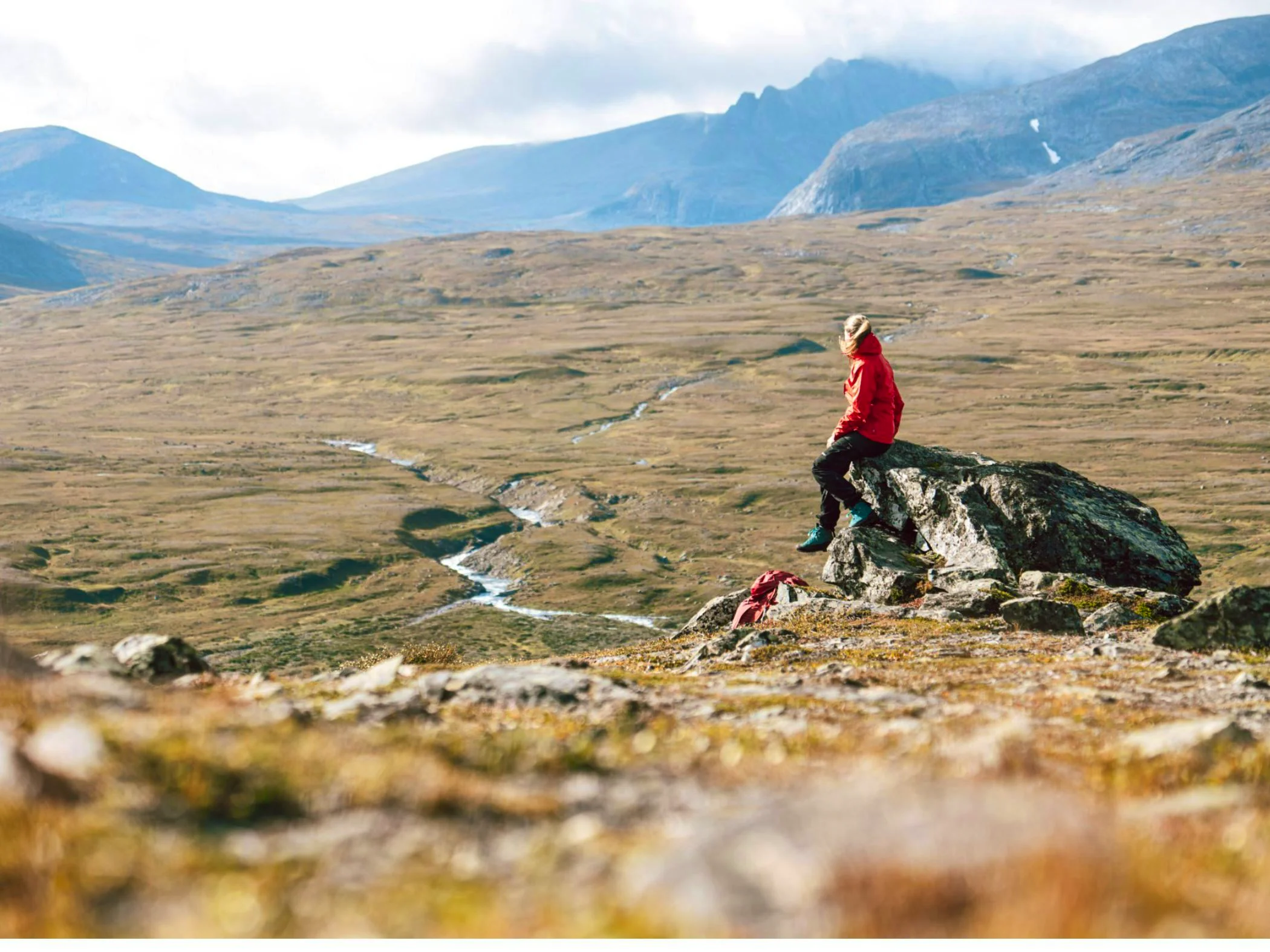 A person in a red jacket sits on a rock, gazing out over a mountain landscape with a winding stream and hills in the background.