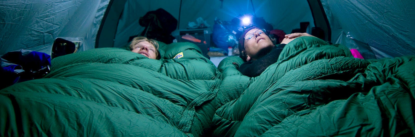 Two people with head torches in separate sleeping bags in a tent.