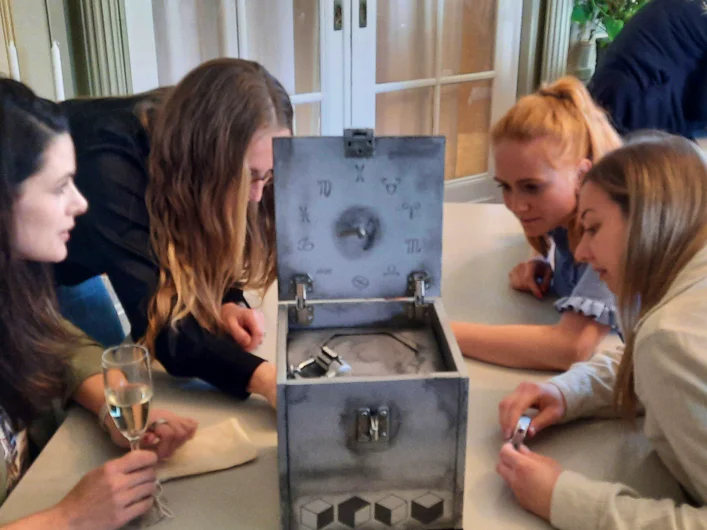 Four people sit around an open box with symbols on the lid, exploring its contents together at a table.