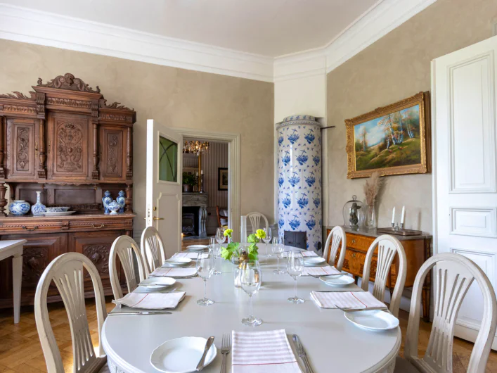 Dining room with a set table, white chairs, a tiled stove, and classic furnishings in a bright, elegant setting.