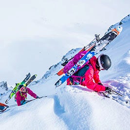 Two skiers dressed in pink, carrying their skis on their backs, make their way up a snow-covered peak.