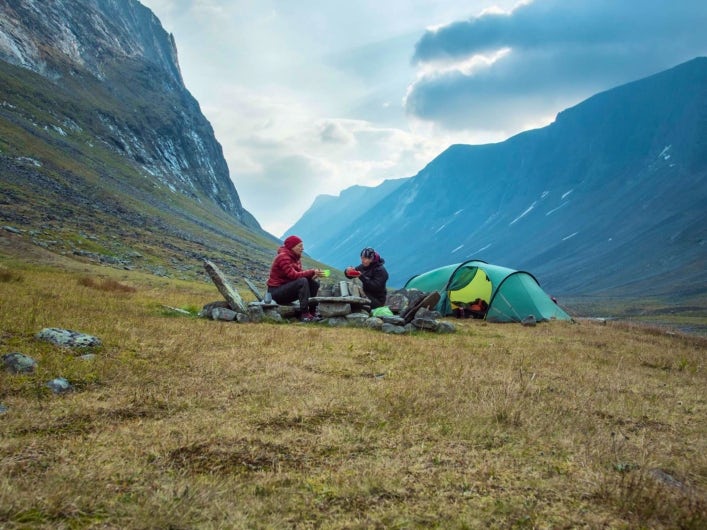 Två personer sitter bredvid ett tält och äter mat i fjällen.