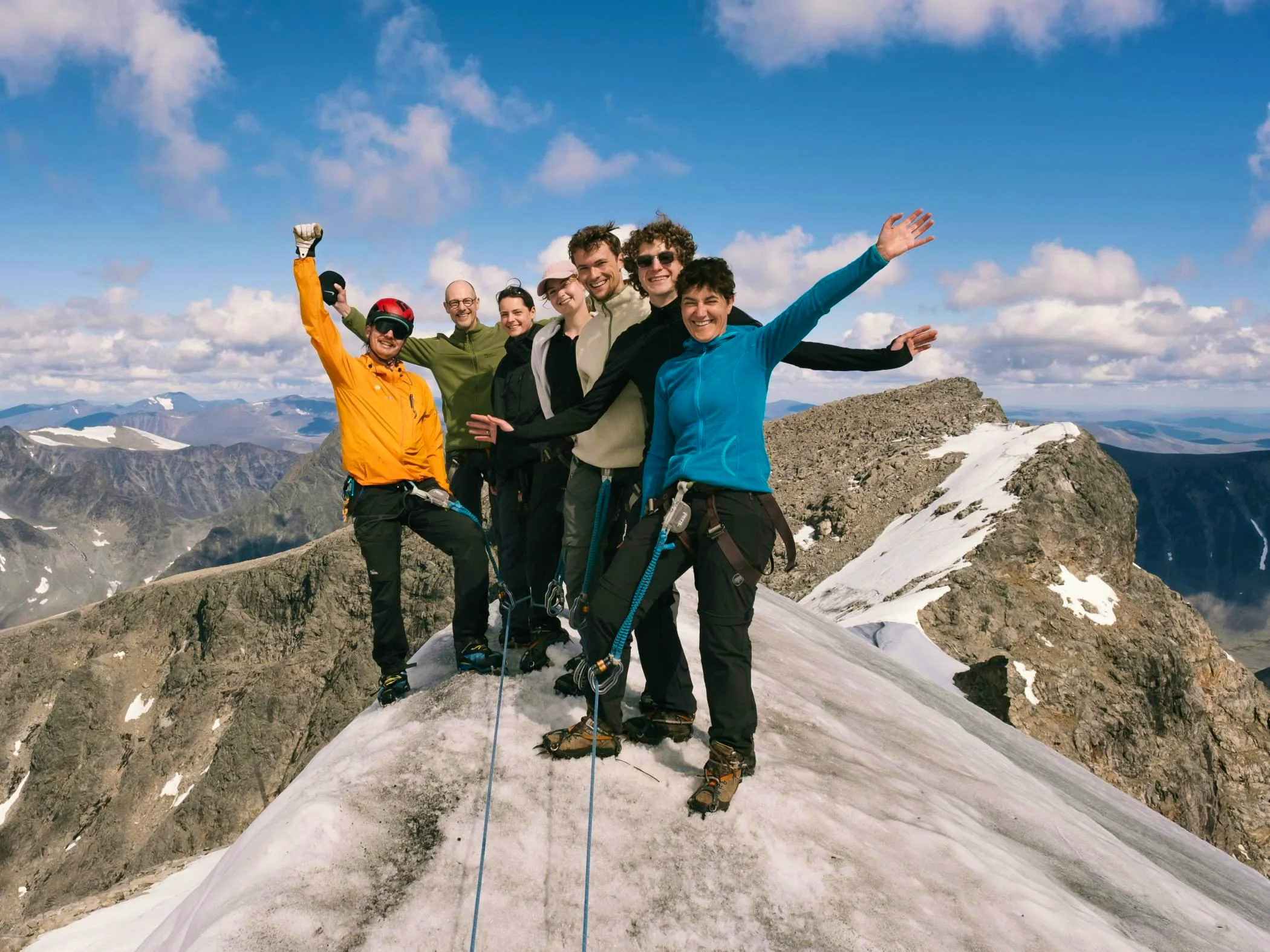 A group of people stands on Kebnekaise's southern peak, equipped with climbing gear, celebrating with a stunning mountain view under a clear blue sky.