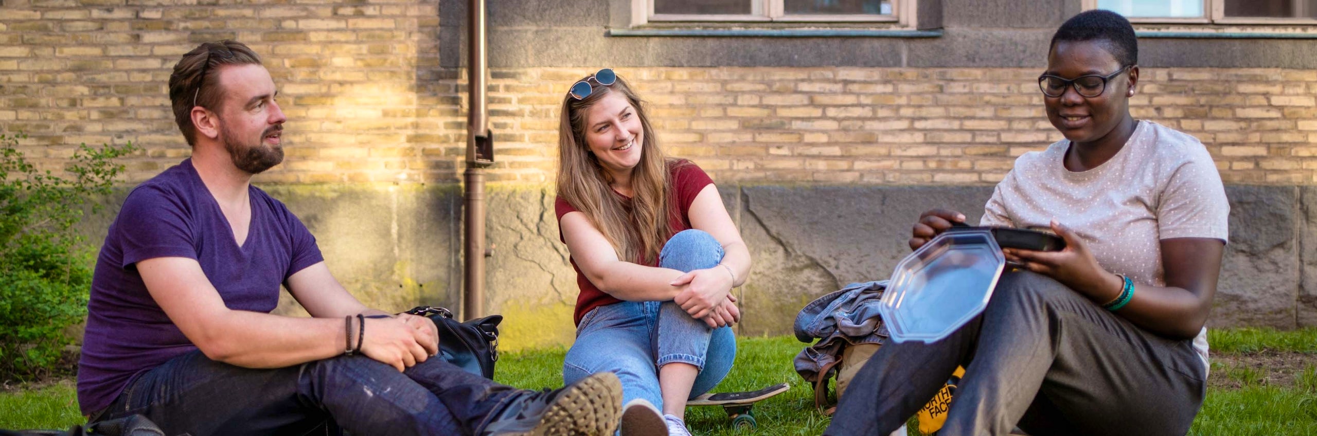 Three people sit relaxed on the grass outside a building, talking and laughing together on a sunny day.