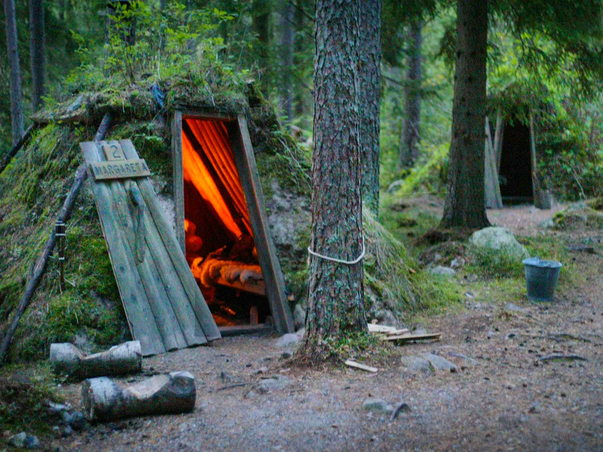 A small turf hut in the forest with warm light glowing from the open doorway.