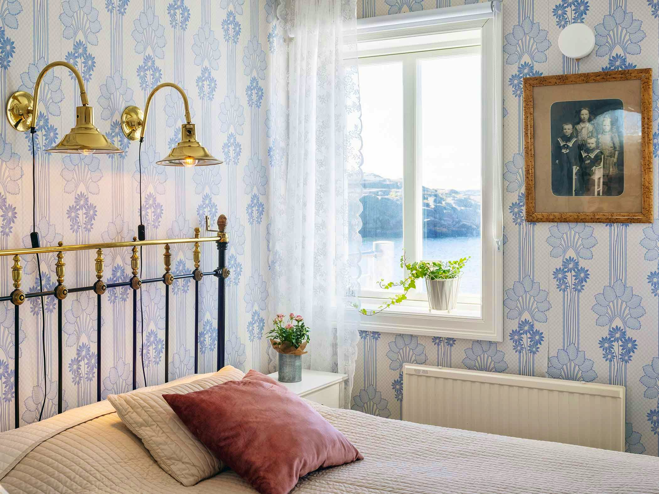 A bright, vintage-style bedroom with floral wallpaper, brass lamps, and a sea view through the window.