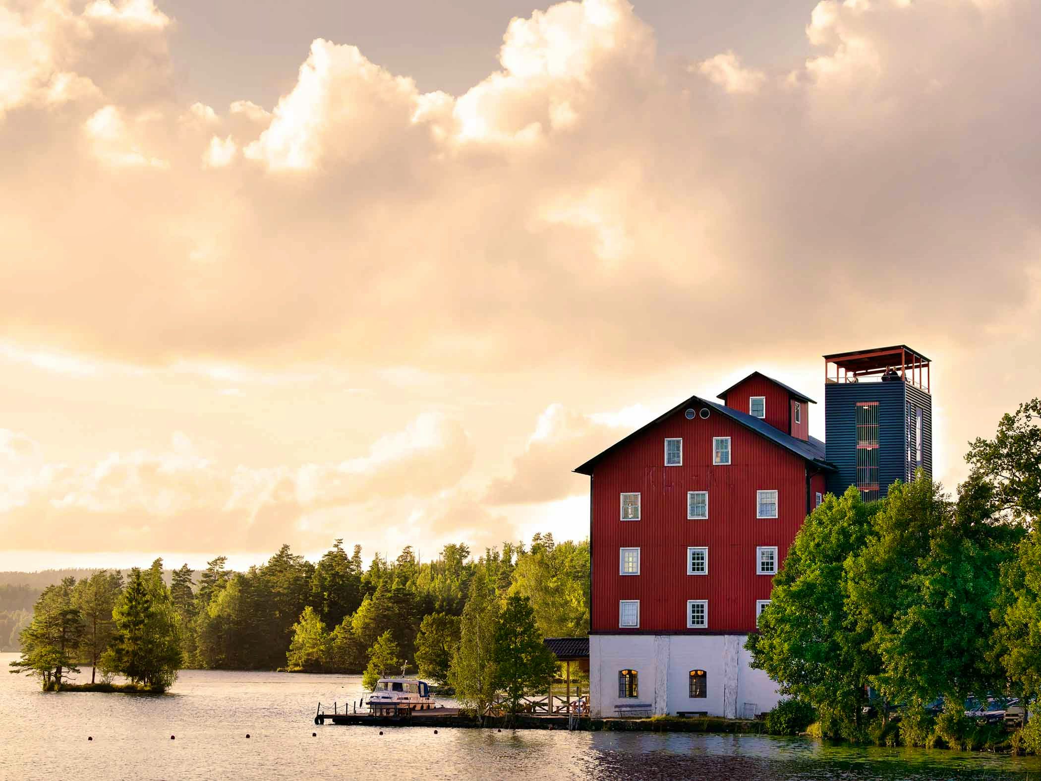 A red wooden building and converted silo stand by a calm lake surrounded by forest, with warm evening light reflecting on the water.