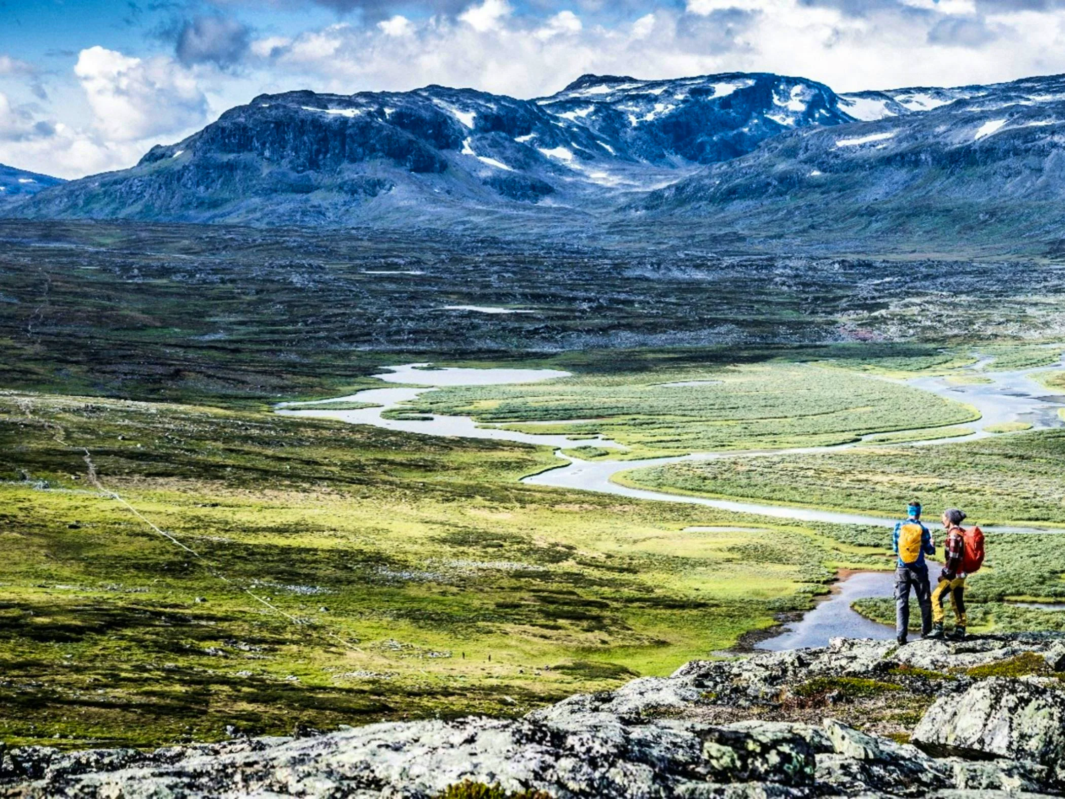 Two hikers with backpacks gaze out over a vast mountain landscape with winding waterways and towering peaks in the background.