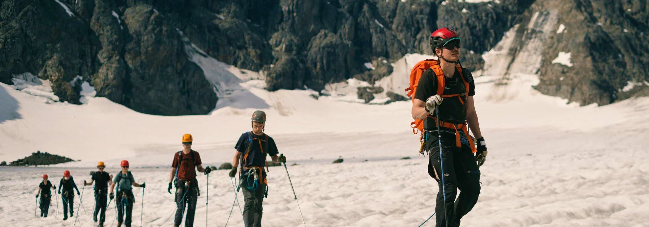 A group of climbers with helmets and harnesses hike on a glacier, led by a guide. Tall cliffs and snow are visible in the background.