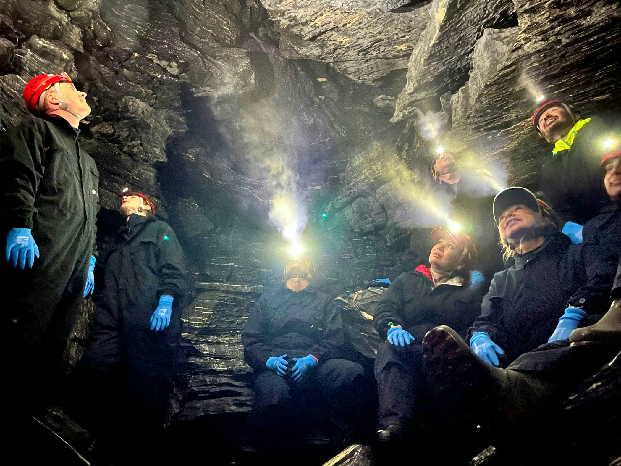 A group of people wearing coveralls, helmets with headlamps, and gloves are sitting and standing inside a dark cave with shiny rock walls. The headlamp beams illuminate the narrow cave.