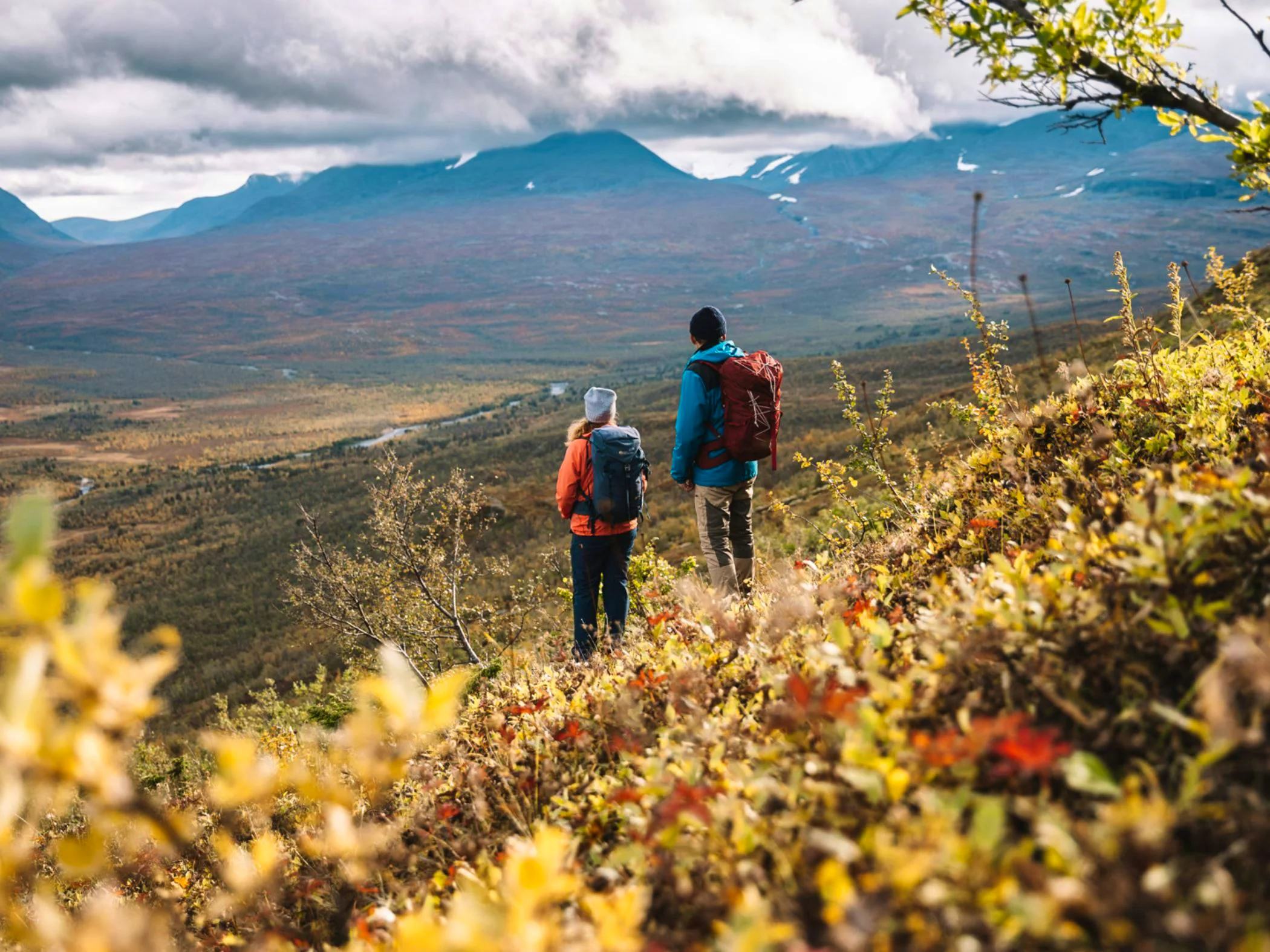 Two hikers with backpacks stand on a slope covered in autumn colors, looking out over a valley with mountains in the background. The sky is cloudy, and the landscape stretches out below them.