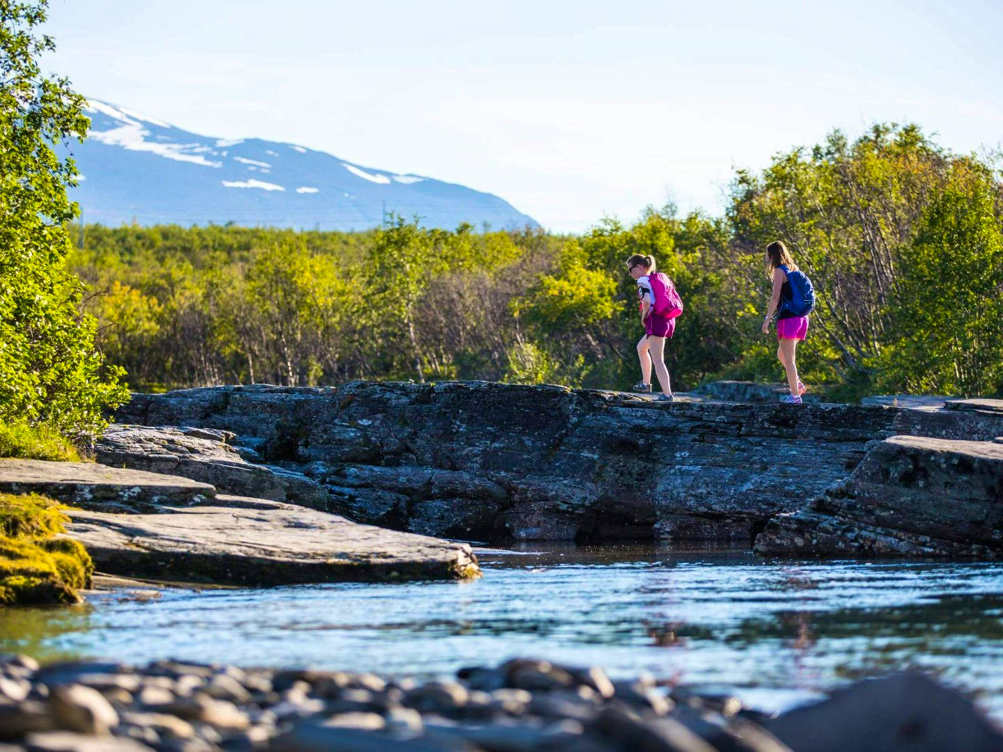 Two people are hiking over rocks near a stream with mountains and forest in the background.