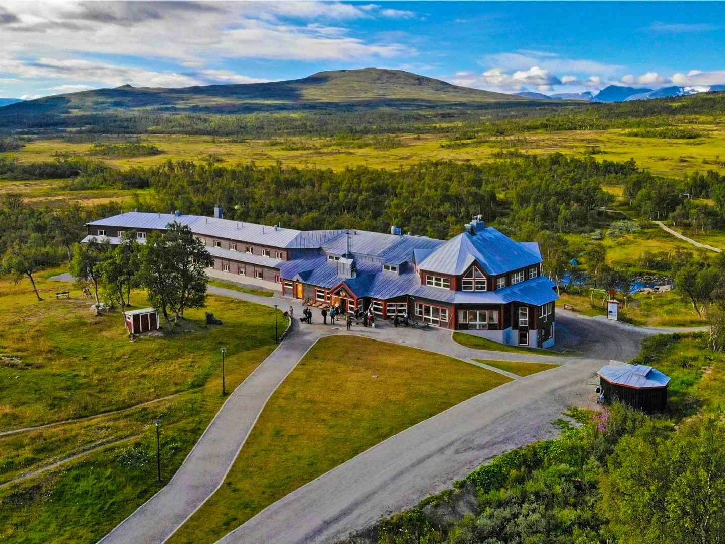 Aerial view of STF Storulvån Mountain Station, a large building with red wooden facades and white details, surrounded by lush mountain scenery. In the background, expansive mountains and a blue sky with clouds are visible. A paved road leads up to the mountain station.