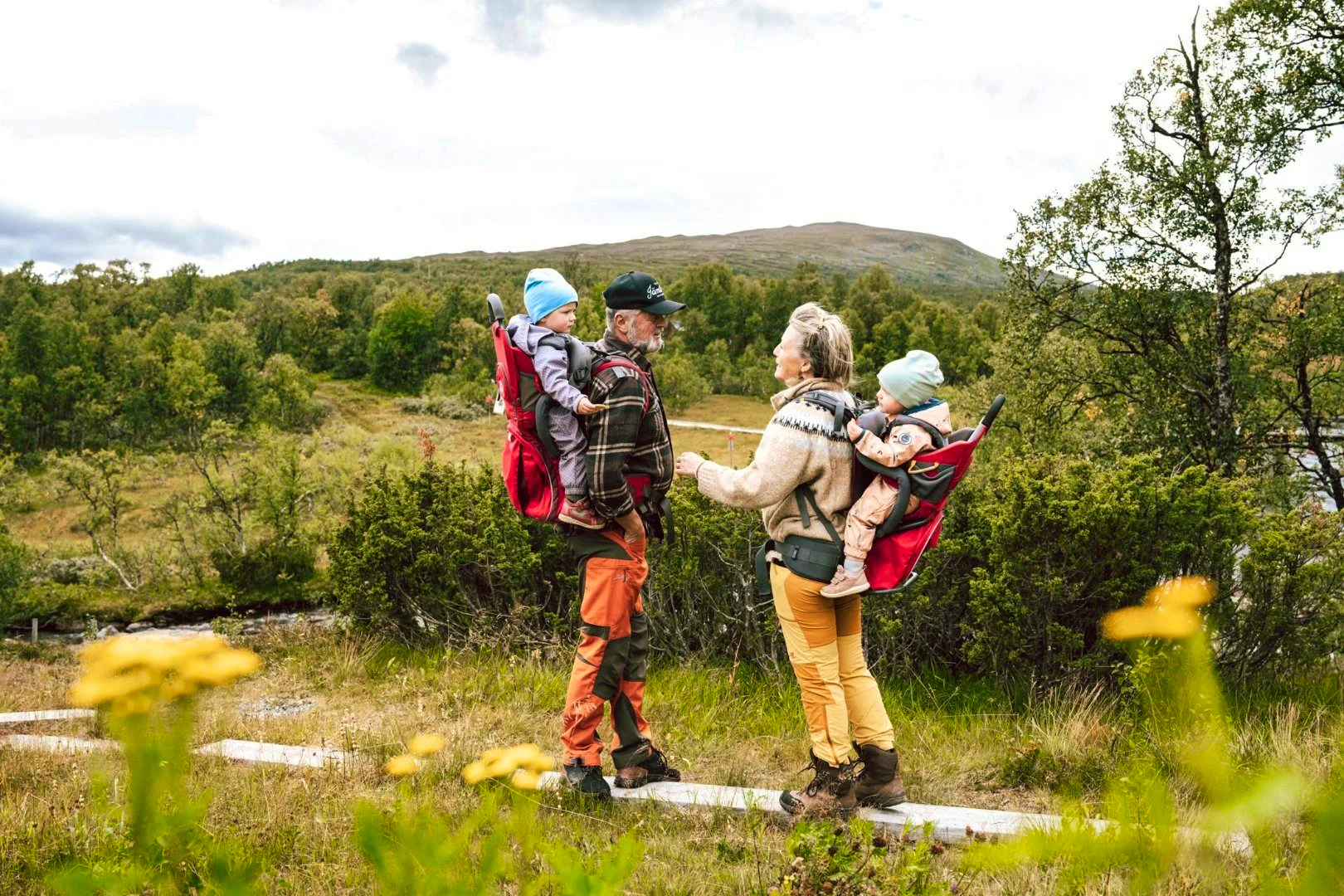 Two adults each carry a child in a child carrier on their backs. They are standing on a trail surrounded by greenery, bushes, and mountains in the background. The image conveys a warm and tranquil feeling, with touches of yellow flowers in the foreground.