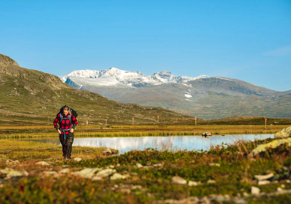 Hiker walking beside a mountain lake with snow-covered peaks in the background.
