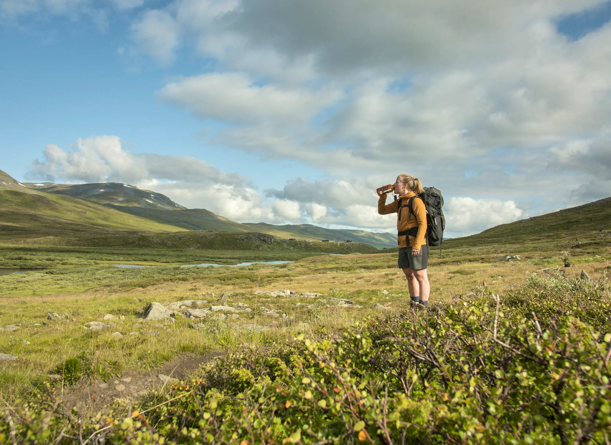 Hiker with a backpack drinking from a bottle in an open mountain landscape with rolling hills and a river under a partly cloudy sky.