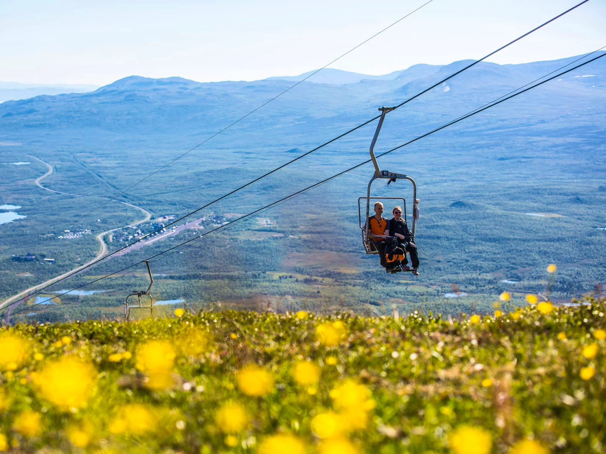 Two people are riding a chairlift over Mount Nuolja with yellow flowers in the foreground.