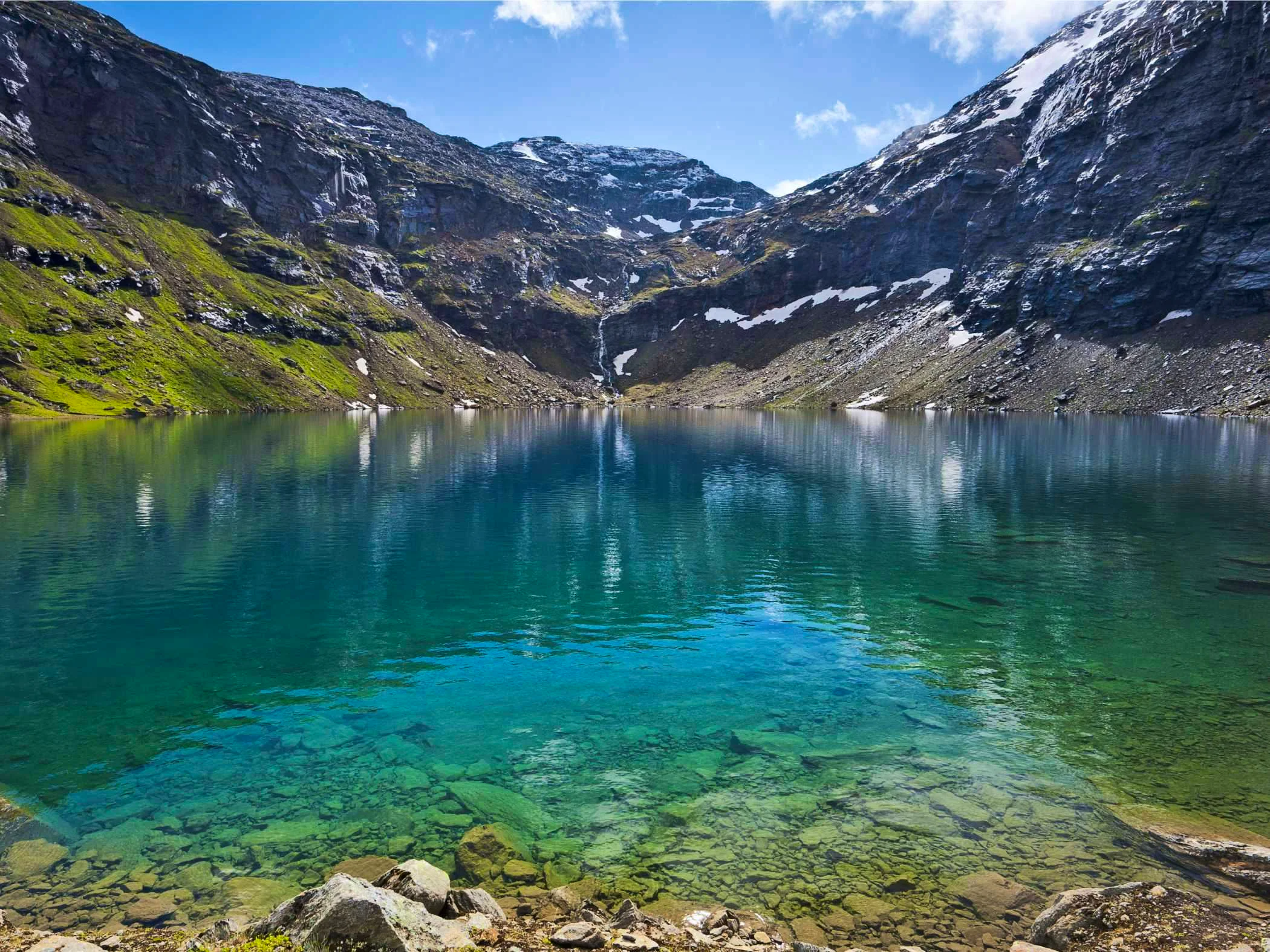 Clear mountain lake surrounded by rocky peaks and patches of snow.