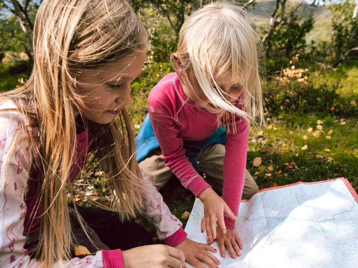 Two children are sitting outdoors in nature, looking at a map together.