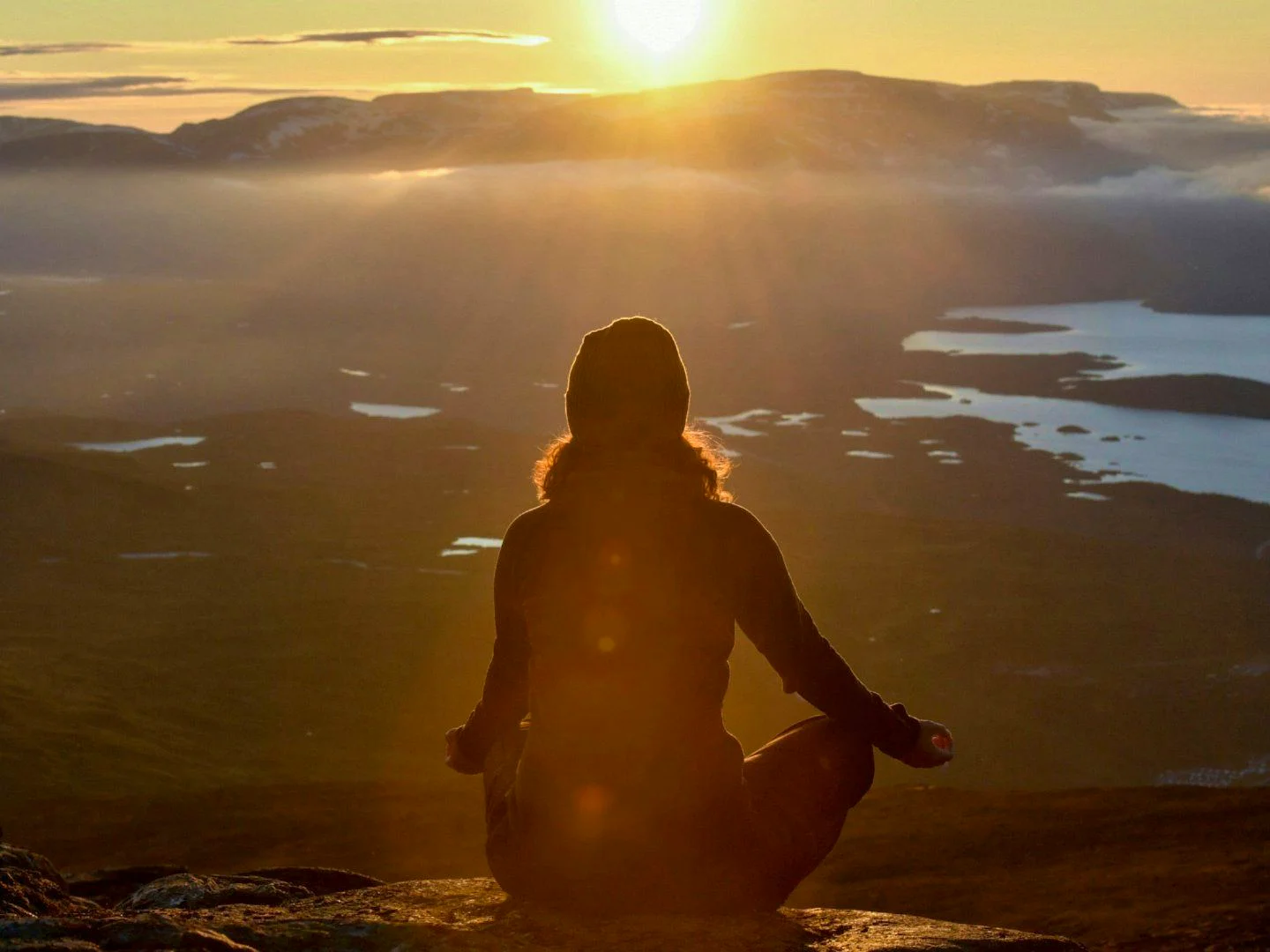 A person sits in a meditation pose on a mountaintop at sunset, overlooking lakes and mountains in the distance.