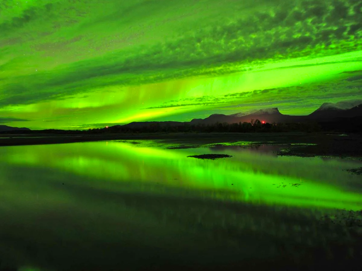 Northern lights reflected in the calm waters of Lake Torneträsk with mountain silhouettes in the background.