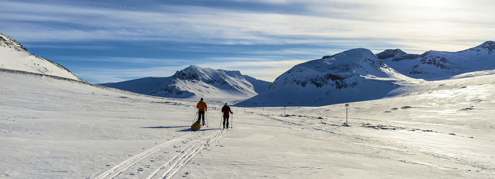 Two skiers pulling pulks across a snow-covered mountain plain, surrounded by high peaks under a clear blue sky.